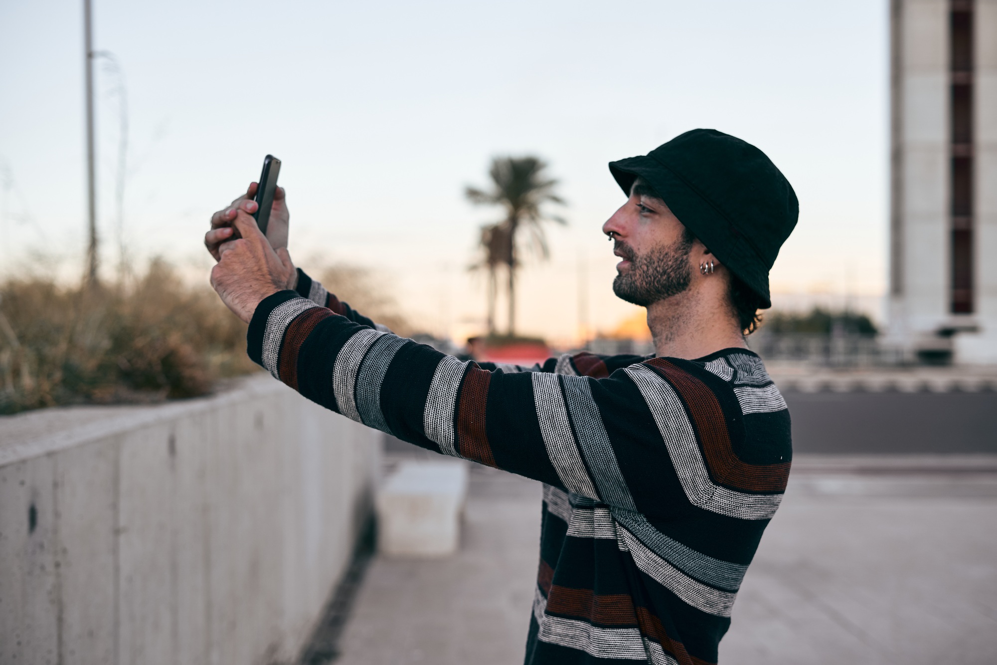 Caucasian guy in profile with nose piercing in striped sweater taking a selfie in the city